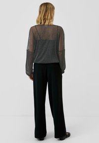 Vrouw met schouderlang blond haar, gekleed in een doorschijnende glinsterende zwarte lange mouw top over een zwarte camisole en zwarte wijde broek, staande.