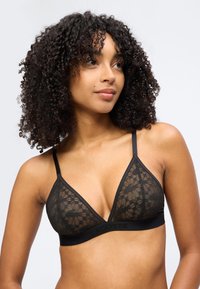 Femme aux cheveux bouclés portant un bralette en dentelle noire à fines bretelles, regardant légèrement vers sa droite sur un fond clair uni.