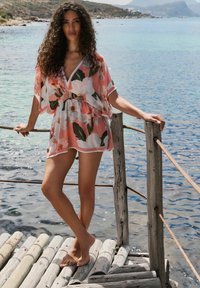 Rózsaszín virágmintás takaró, mély V-dekoltázzsal, laza szabással és rakott derékkal. Könnyű anyag fehér díszítéssel. Mezítláb viselik egy fából készült stégen.