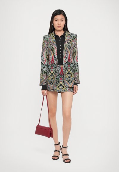 Patroon blazer- en shortset in grijs, rood, groen en gele paisleydesign, gecombineerd met een zwarte polo en een rode handtas. Zwarte sandalen met banden.