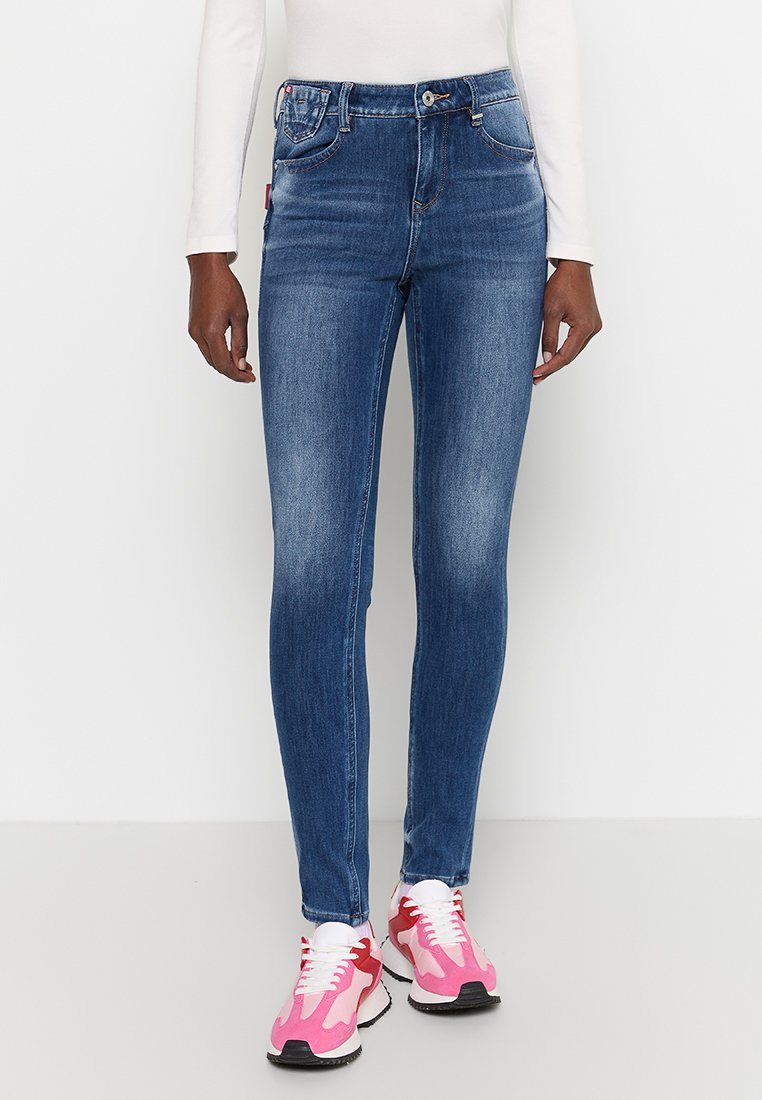 Jeans skinny blu in denim con dettagli di whiskering, tasche anteriori e chiusura a bottone, abbinati a sneakers sportive rosa e bianche.