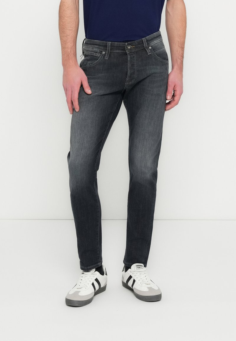 Mies, jolla on haalistuneet mustat kapeat farkut, valkoiset lenkkarit mustilla raidoilla ja tummansininen paita, seisoo valkoista taustaa vasten.