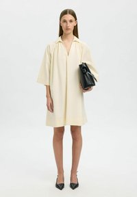 Vestido corto crema con cuello y hombros caídos, combinado con un bolso negro y tacones negros sofisticados, situado sobre un fondo neutro.