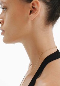 Un collar de cadena dorada cuelga alrededor del cuello, combinado con una blusa negra y texturizada. La piel tiene una complexión suave con pequeñas pecas sutiles.