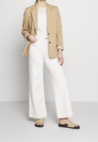 Béžový blazer, bílý top bez rukávů, bílé široké kalhoty a černé sandály s světle béžovými akcenty. Hladká tkanina a strukturovaný design.