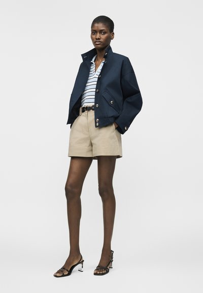 Γυναίκα με κοντό ναυτικό σακάκι πάνω από λευκό και ναυτικό ριγέ πουκάμισο, μπεζ σορτς με ζώνη και μαύρα πέδιλα με τακούνια και λουράκια.