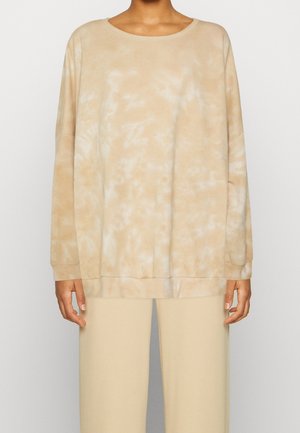 Personne portant un sweat-shirt à manches longues tie-dye beige et blanc avec un pantalon beige assorti, debout devant un fond clair uni.