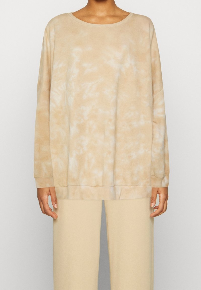 Personne portant un sweat-shirt à manches longues tie-dye beige et blanc avec un pantalon beige assorti, debout devant un fond clair uni.