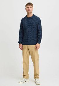 Marineblauwe cable-knit trui met geribbelde manchetten en onderrand, gecombineerd met beige chino's en witte sneakers. Model staat voor een neutrale achtergrond.