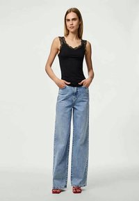 Svart tanktopp med blonder, kombinert med lyseblå vide bukser. Modellen har på seg røde sandaler og står foran en ensfarget bakgrunn.