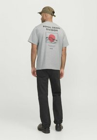Camiseta gris de manga corta con un diseño gráfico que presenta un círculo rojo y montañas, etiquetada "DIVISIÓN DENIM REAL". Pantalones negros y gorra.