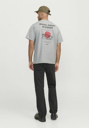 Grijze T-shirt met korte mouwen en een grafisch ontwerp met een rode cirkel en bergen, met het label "ROYAL DENIM DIVISION." Zwarte broek en pet.