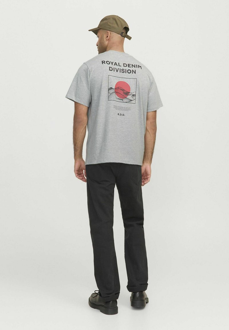 Camiseta gris de manga corta con un diseño gráfico que presenta un círculo rojo y montañas, etiquetada "DIVISIÓN DENIM REAL". Pantalones negros y gorra.