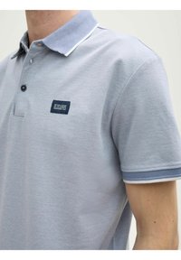 Grå pikétröja med texturerad tyg, tvåknappsslutning och kontrasterande krage. Inkluderar en liten marinblå logotyp på bröstet.