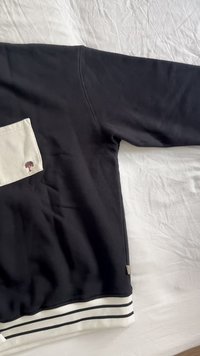 Melna treniņjakas ar krēmkrāsas kabatu. Aprīkots ar baltām un melnām svītrām jostasvietā. Mīksta tekstūra, ikdienišķs dizains, ar mazu logotipu uz kabatas.