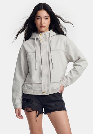 Jeune femme aux longs cheveux foncés portant une veste à capuche gris clair et un short en jean noir déchiré, debout avec une main sur la hanche.