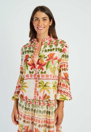 Donna sorridente, che indossa una camicetta chiara e una gonna con motivi floreali e di palme colorati, in piedi davanti a uno sfondo uniforme.