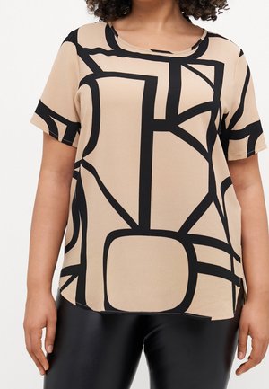 Kvinde iført beige kortærmet top med abstrakte sorte geometriske mønstre og sorte læderbukser, armene afslappet ned langs siderne.