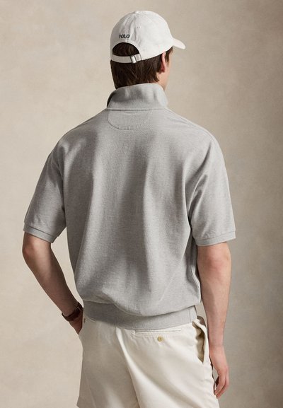 Άνδρας που φορά ανοιχτό γκρι πουλόβερ με κοντό μανίκι και ψηλό γιακά και λευκό καπέλο, στέκεται με το ένα χέρι στην τσέπη σε απλό φόντο.