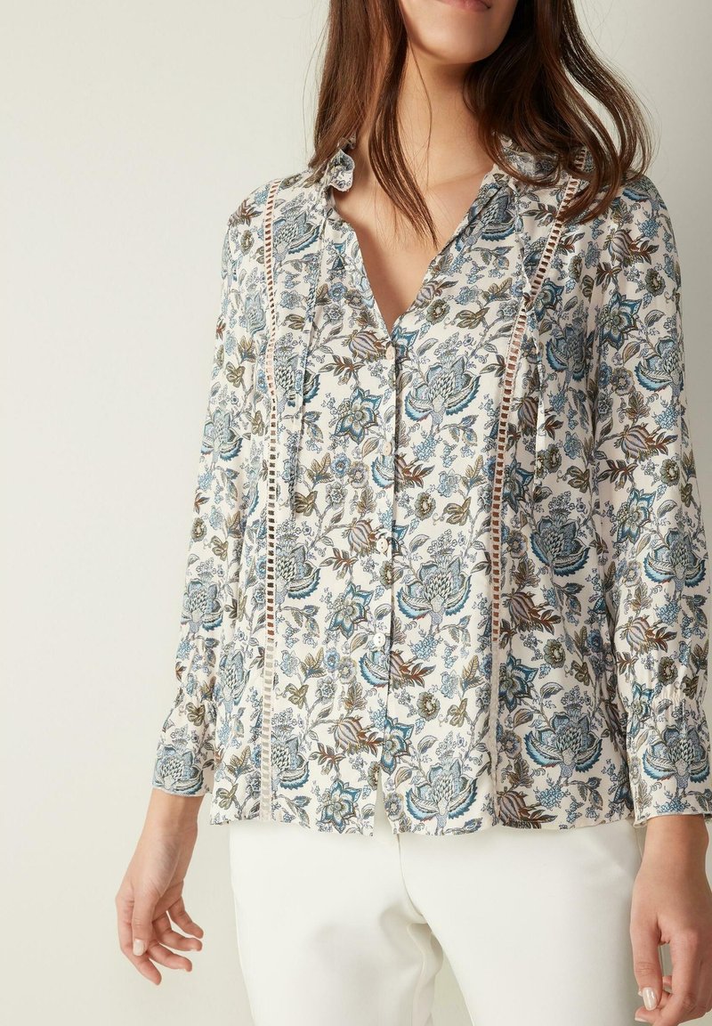 Femme portant un chemisier blanc à manches longues à motifs floraux avec des détails en dentelle, associé à un pantalon blanc sur un fond uni.