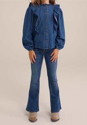 Enfant portant un chemisier en jean bleu à boutons avec volants, un jean évasé bleu et des bottines beiges, debout sur fond neutre.