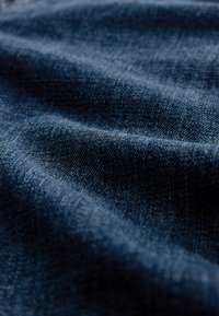 Gros plan sur un tissu en denim bleu foncé avec une texture de tissage visible et des plis doux créant des motifs de lumière et d’ombre.