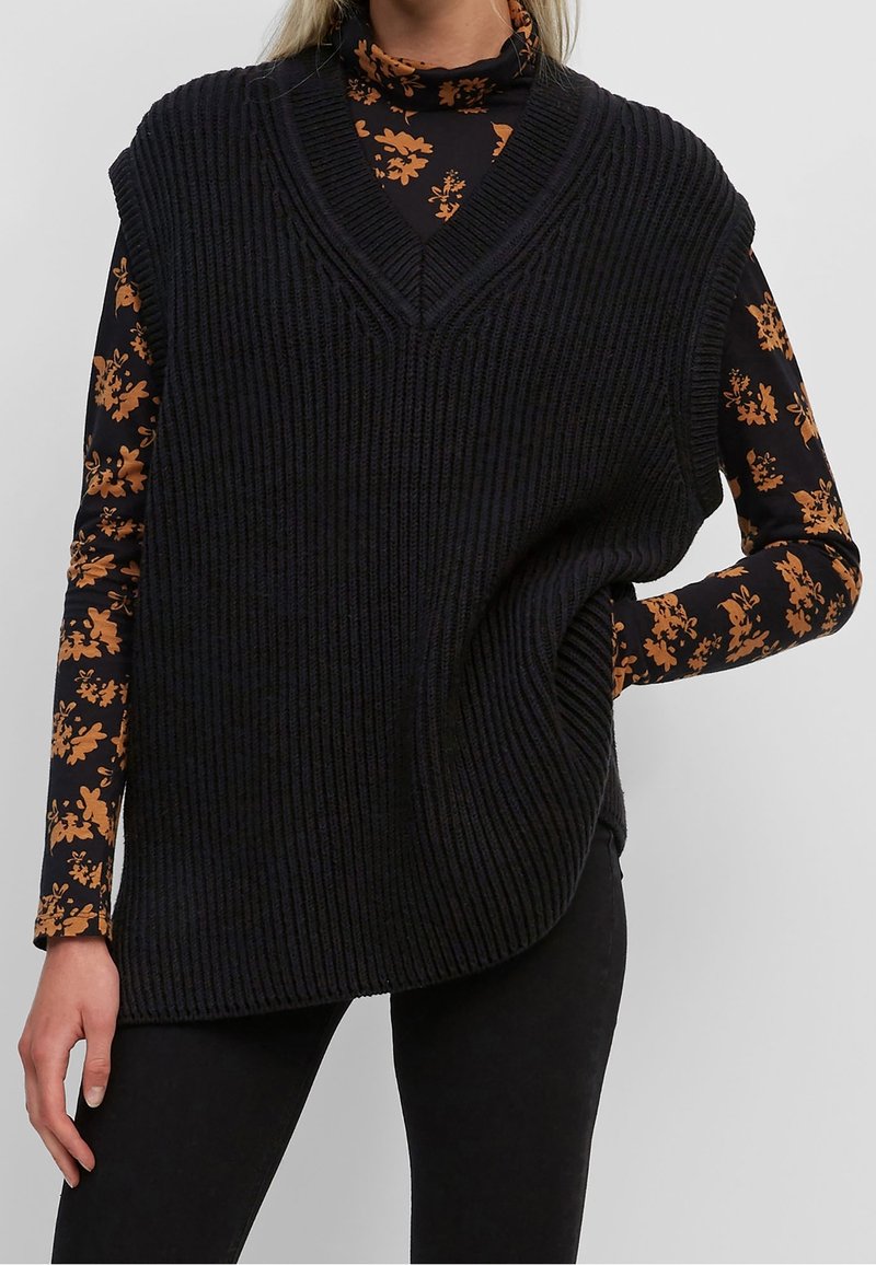 Gilet senza maniche a costine nere con scollatura a V profonda e vestibilità rilassata, indossato sopra un maglione a collo alto con motivi floreali arancioni.
