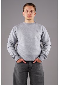 Grauer Sweatshirt mit geripptem Rundhalsausschnitt und Bündchen. Verfügt über ein kleines Logo auf der Brust. Eine dunkelgraue Jeans rundet den Look ab.