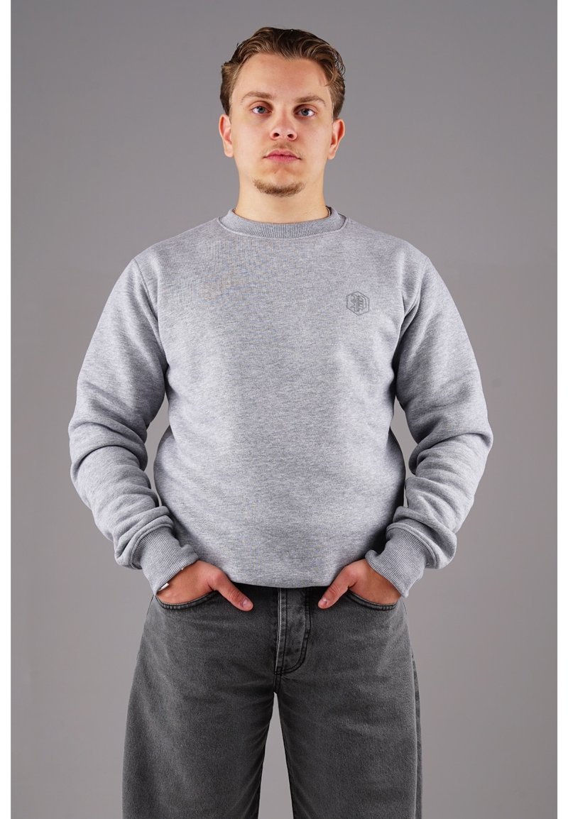 Grauer Sweatshirt mit geripptem Rundhalsausschnitt und Bündchen. Verfügt über ein kleines Logo auf der Brust. Eine dunkelgraue Jeans rundet den Look ab.