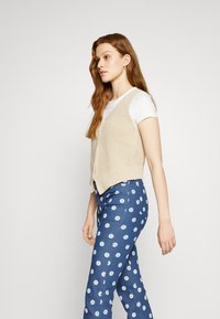 Colete bege às malhas sem mangas sobre uma t-shirt branca, combinado com calças azuis estampadas com pequenas flores brancas. Vista lateral, textura suave.