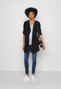 Chaqueta negra de corte largo con mangas enrolladas, blusa de color azul claro, jeans ajustados azules y bambas blancas. Lleva un bolso negro con detalles redondeados.