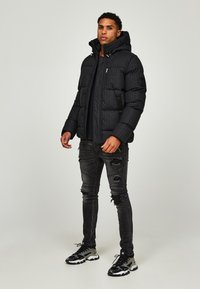 Svart jakke med hette, mønstrete tekstur, glidelås-lukking, kombinert med slitte svarte jeans og svarte sneakers med grå aksenter.