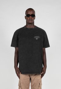 Sort oversized bomulds T-shirt med rund halsudskæring og korte ærmer, prydet med hvid tekst foran. Bæres med beige cargo bukser.