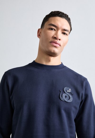 Pullover blu navy con una patch texturizzata a forma di serpente sul lato sinistro del petto, con collo rotondo e polsini a costine.