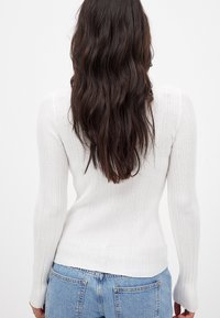 Weißes, geripptes Langarmoberteil mit tailliertem Schnitt. Gerader Saum und leicht ausgestellte Bündchen. Getragen mit blauen Denim-Jeans.