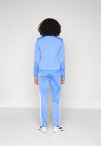 Ανοιχτό μπλε αθλητική φόρμα κατασκευασμένη από απαλό, ελαστικό ύφασμα. Διαθέτει λευκές ρίγες στο πλάι του παντελονιού και ριμπ μανσέτες στο μπουφάν και στο παντελόνι.