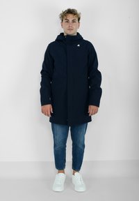 Námornícka vodeodolná parka so zipsom vpredu, nastaviteľnou kapucňou a diskrétnym logom na ľavej hrudi. Skombinovaná so svetlomodrými džínami a bielymi teniskami.