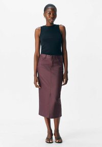 Femme portant un haut sans manches noir, une jupe midi bordeaux taille haute, des sandales noires, se tenant devant un fond blanc uni.