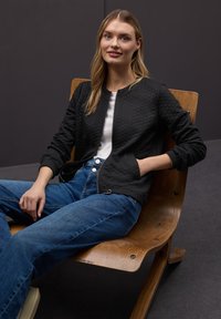 Czarna pikowana kurtka bomberka z okrągłym dekoltem, noszona na białej bluzce, zestawiona z niebieskimi dżinsami. Siedzi na drewnianym krześle.