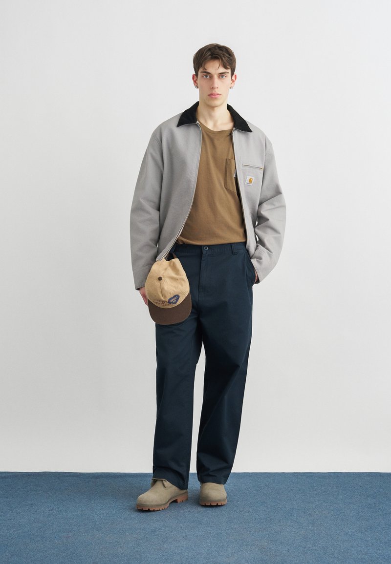 Junger Mann steht, hält eine beige Mütze in der Hand, trägt eine graue Jacke, ein braunes Hemd, marineblaue Hose und beige Schuhe auf blauem Teppich.