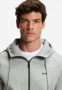 Grijze rits hoodie met verstelbare trekkoorden, voorzien van een zwarte rits en logo op de borst, gemaakt van een zachte, textuurstof.