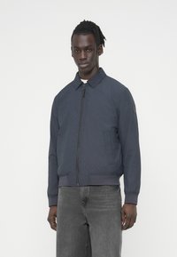 Μπλε ναυτικό μπουφάν bomber με φερμουάρ, ριμπ μανσέτες και ζώνη μέσης, με διακριτική υφή με ραφές. Φορεμένο πάνω από ανοιχτόχρωμο τοπ.