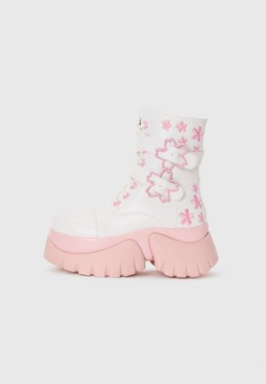 Stivale bianco con punta arrotondata, caratterizzato da accenti floreali rosa e due cinturini rosa. Suola testurizzata con scanalature prominenti.