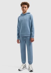 Lichtblauwe katoenen hoodie met een logo aan de voorkant, gecombineerd met bijpassende joggingbroeken en zilveren sportschoenen met zwarte accenten.