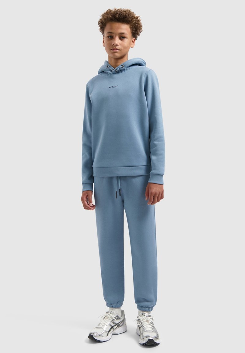 Lichtblauwe katoenen hoodie met een logo aan de voorkant, gecombineerd met bijpassende joggingbroeken en zilveren sportschoenen met zwarte accenten.