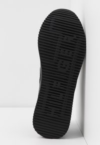 Semelle de chaussure en caoutchouc noir avec un motif texturé et rainuré et le nom de la marque en relief sur la surface, posée sur un fond blanc.