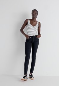 Camiseta sin mangas blanca con rayas negras, combinada con jeans ajustados negros y zapatillas negras con acentos blancos y suela de goma. Pose de pie.