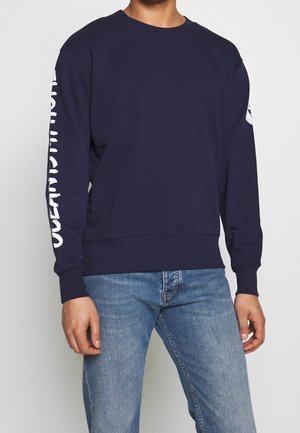 Mand iført navyblå sweatshirt med hvid tekst på højre ærme og lyseblå jeans, stående mod en neutral baggrund.