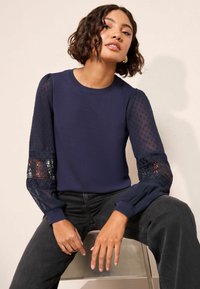 Tumesinine kampsun tekstuurse koo kehaosaga ja läbipaistvate täpiliste varrukatega. Mansettidel pitsdetailid. Modell istub ja kannab musti pükse.