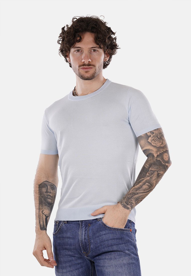 Maglietta azzurra chiara a maniche corte con scollatura rotonda, texture a coste e design aderente, abbinata a jeans scuri e con tatuaggi visibili.